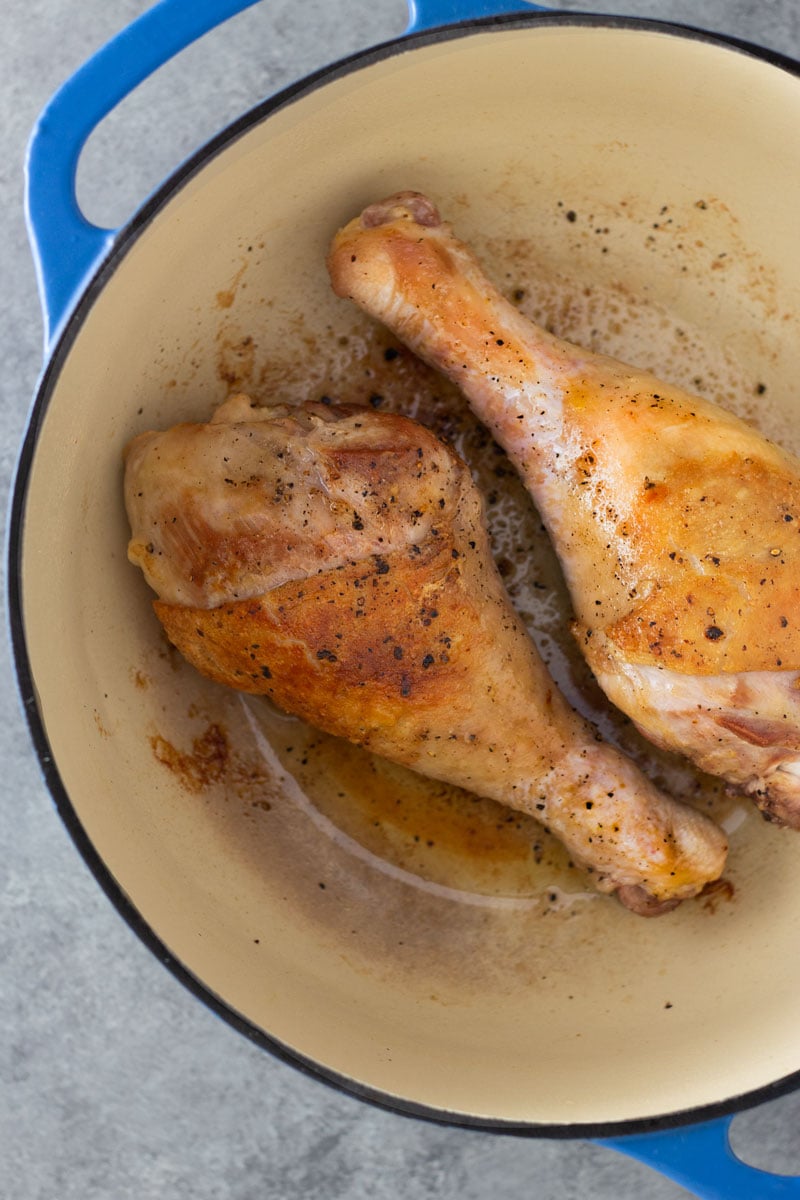 Overhead view on a blue dutch oven filled with two browned turkey legs on a light grey, textured surface. 