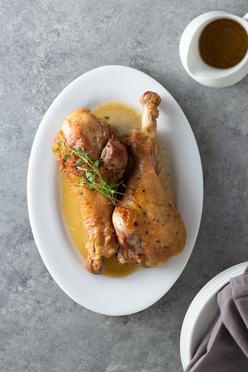Overhead view of two Apple Cider Braised Turkey Legs on a serving platter topped with fresh thyme surrounded by a pitcher of apple cider jus and plates and napkins on a light grey, textured surface. 
