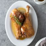 Overhead view of two Apple Cider Braised Turkey Legs on a serving platter topped with fresh thyme surrounded by a pitcher of apple cider jus and plates and napkins on a light grey, textured surface.