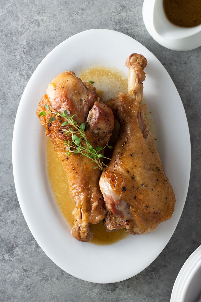 Overhead view of two Apple Cider Braised Turkey Legs on a serving platter topped with fresh thyme surrounded by a pitcher of apple cider jus and plates on a light grey, textured surface.