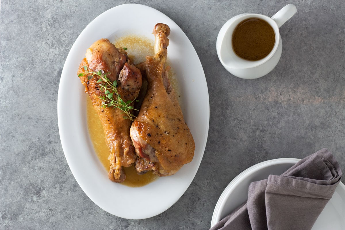 Overhead view of two Apple Cider Braised Turkey Legs on a serving platter topped with fresh thyme surrounded by a pitcher of apple cider jus and plates and napkins on a light grey, textured surface.