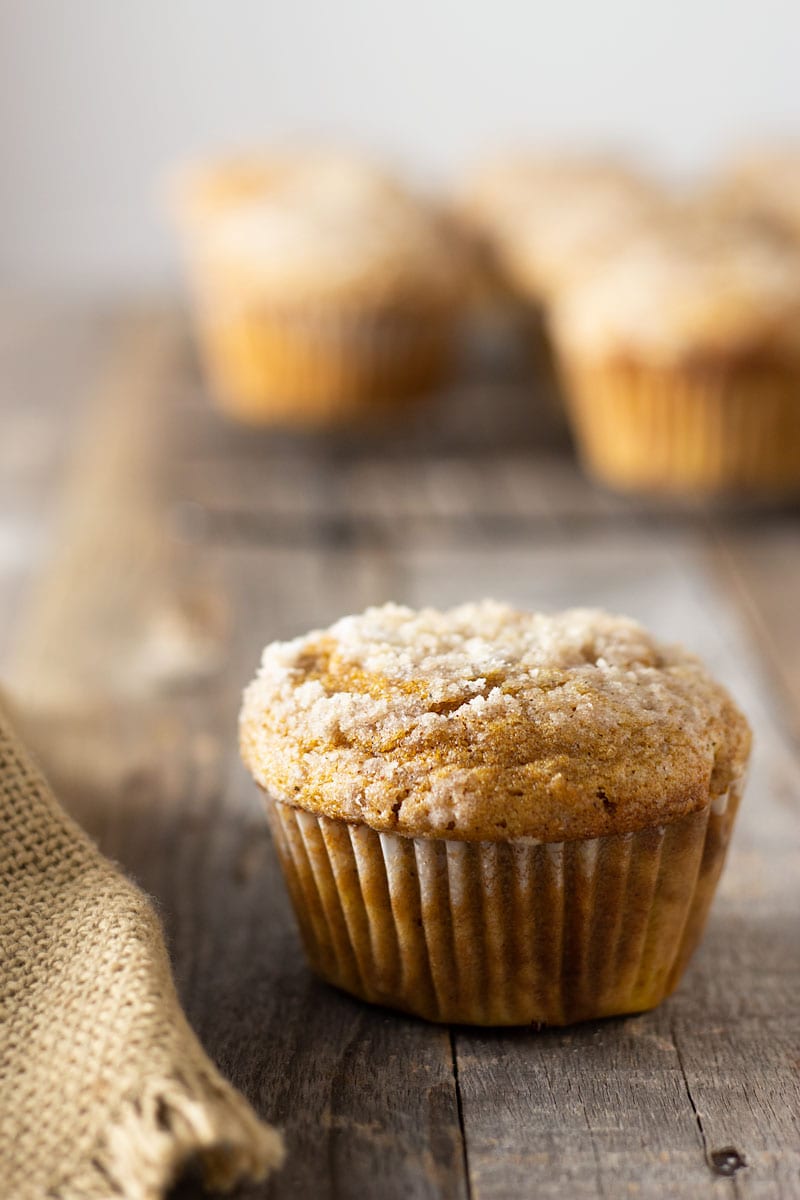Straight on shot of a Pumpkin Cream Cheese Streusel Muffin on a grey wood surface surrounded by a tan cloth with muffins on a cooling rack in the background.