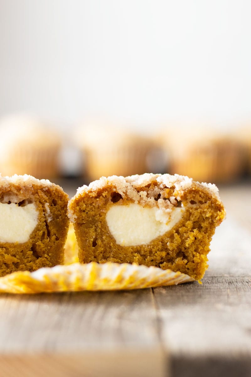 Straight on shot of a Pumpkin Cream Cheese Streusel Muffin cut open to reveal the cream cheese center on a grey wood surface with muffins in the background.