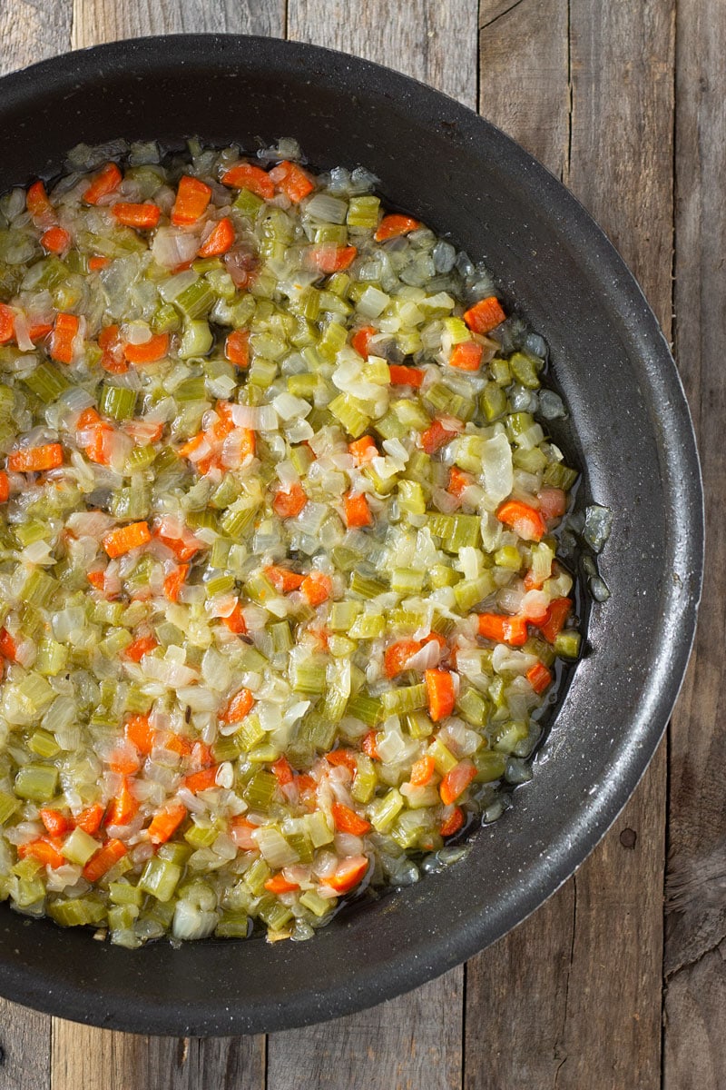 Process shot of cooking celery, carrots and onion in a pan for Sausage Chestnut Stuffing on a rustic wood surface.