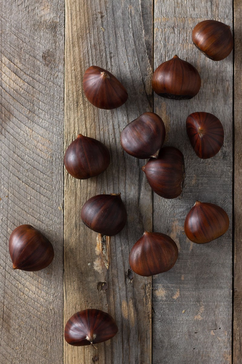 Ingredient shot of raw chestnuts scattered on a rustic wood surface for Chestnut Sausage Stuffing.