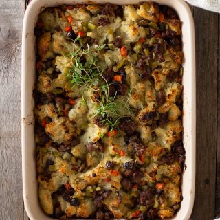 Overhead view of hands in a white sweater holding a red baking tray of Sausage Chestnut Stuffing topped with fresh thyme on a grey wood surface.