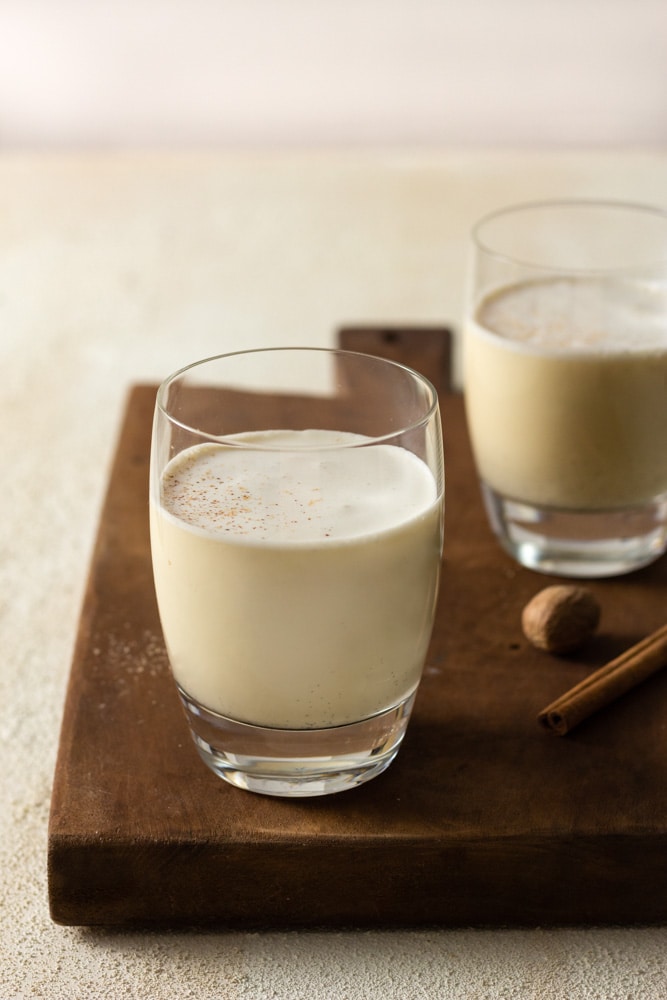 ¾ angled shot of two glasses of vanilla bean eggnog topped with grated nutmeg and cinnamon on a wood cutting board on a textured off white surface.