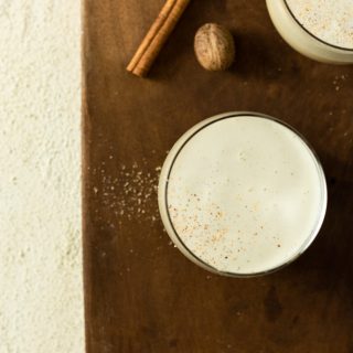 Overhead shot of two glasses of eggnog topped with grated nutmeg and cinnamon on a wood cutting board on a textured off white surface.