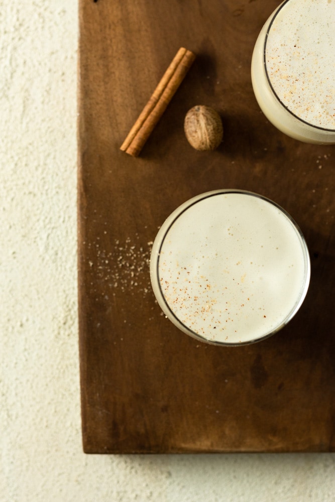 Overhead shot of two glasses of vanilla bean eggnog topped with grated nutmeg and cinnamon on a wood cutting board on a textured off white surface.