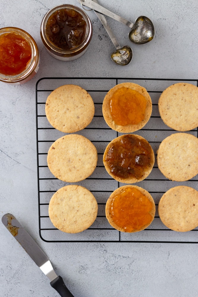 Overhead shot of hazelnut cookies on a cooling rack being filled with apricot and fig jams surrounded by jars of jam, measuring spoons and an offset spatula on a light blue grey surface.