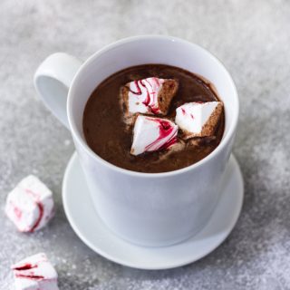 3/4 angled view of a white mug of hot chocolate with homemade peppermint marshmallows on a saucer next to two marshmallows on a light grey surface dusted with powdered sugar.