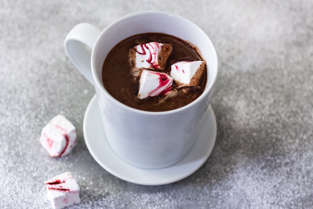 ¾ angled view of a white mug of hot chocolate with homemade peppermint marshmallows on a saucer next to two marshmallows on a light grey surface dusted with powdered sugar.