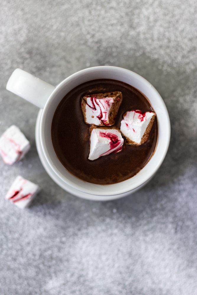 Overhead view of a white mug of hot chocolate with homemade peppermint marshmallows on a saucer next to two marshmallows on a light grey surface dusted with powdered sugar.