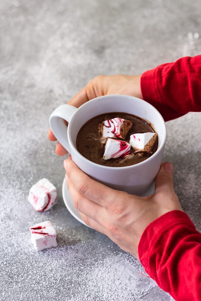 ¾ angled view of hands holding a white mug of hot chocolate with homemade peppermint marshmallows on a saucer next to two marshmallows on a light grey surface dusted with powdered sugar.