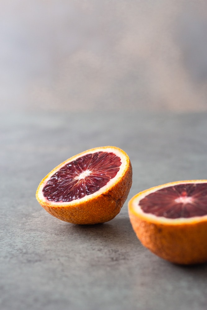 Straight on view of two halves of a blood orange on a light grey textured surface with a blue grey background. 