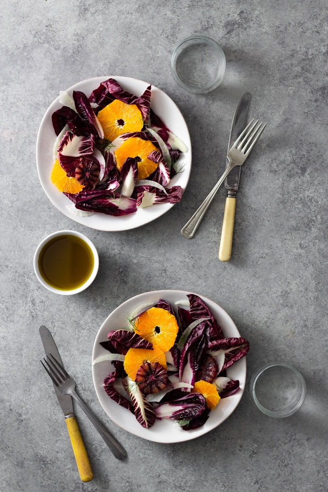 Overhead view of two plates of Orange and Fennel Salad with Radicchio, blood orange and fennel fronds surrounded by glasses of water, forks, knives, and a small bowl of dressing on a light grey, textured surface. 