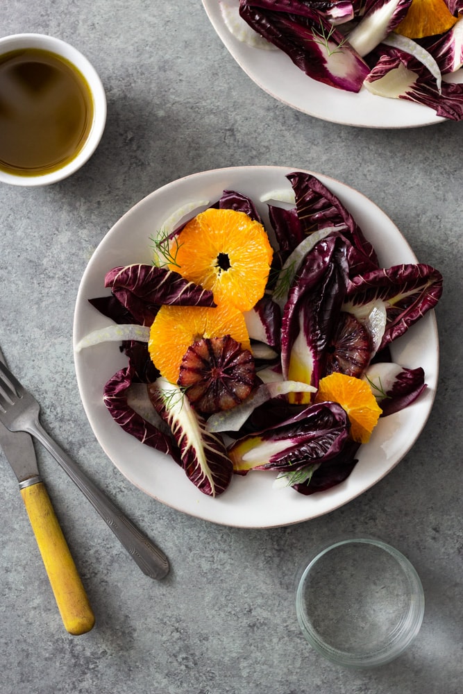 Overhead view of an Orange and Fennel Salad with Radicchio, blood orange, fennel fronds and a sherry vinaigrette on a white plate surrounded by a small bowl of dressing, another plate of salad, a fork, a knife and a glass of water on a grey, textured surface. 