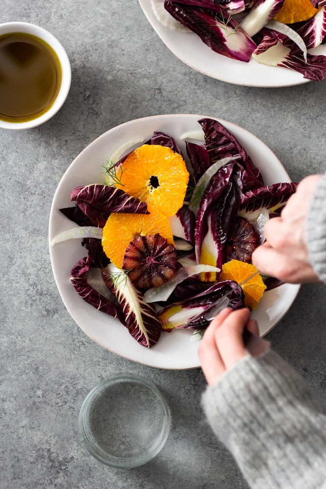Overhead view of hands cutting into an orange and fennel salad with radicchio, blood orange and fennel fronds next to another bowl of salad, a small bowl of sherry vinaigrette and a glass of water on a light grey textured surface.