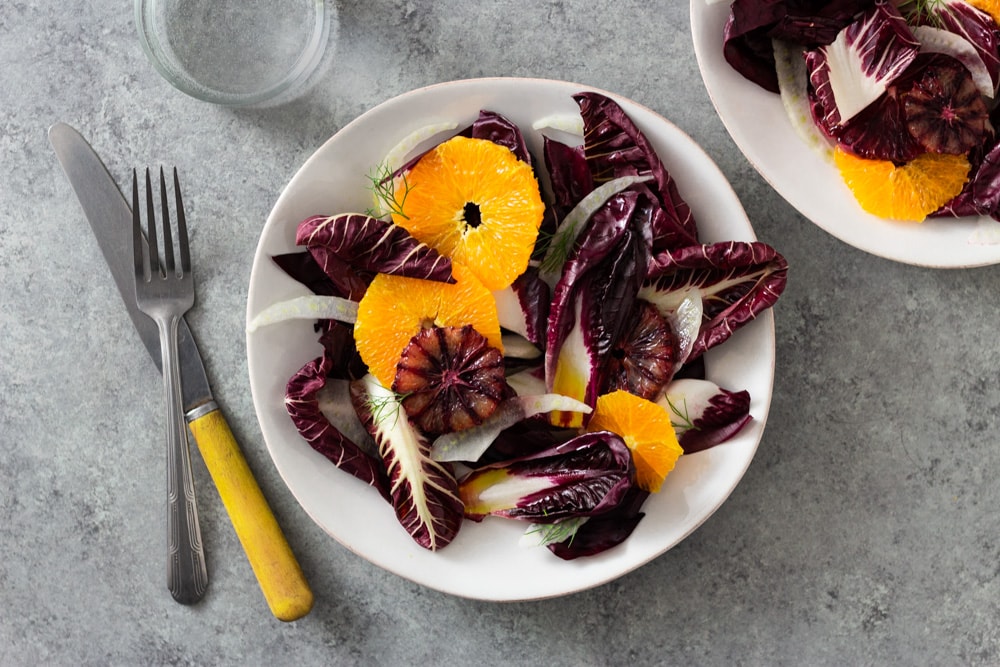 Overhead view of an Orange and Fennel Salad with Radicchio, blood orange, fennel fronds and a sherry vinaigrette on a white plate surrounded by another plate of salad, a fork, a knife and a glass of water on a grey, textured surface. 