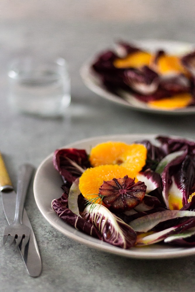 ¾ angled view of an Orange and Fennel Salad with Radicchio, blood orange, fennel fronds and a sherry vinaigrette on a white plate next to a fork and a knife with a glass of water and another salad in the background on a grey, textured surface. 