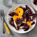 Overhead view of an Orange and Fennel Salad with Radicchio, blood orange, fennel fronds and a sherry vinaigrette on a white plate surrounded by another plate of salad, a fork, a knife and a glass of water on a grey, textured surface.