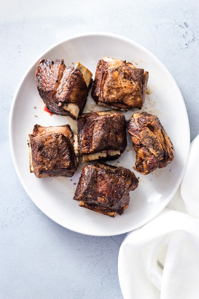 Overhead shot of a white plate with browned beef short ribs next to a white linen napkin on a light blue surface. 