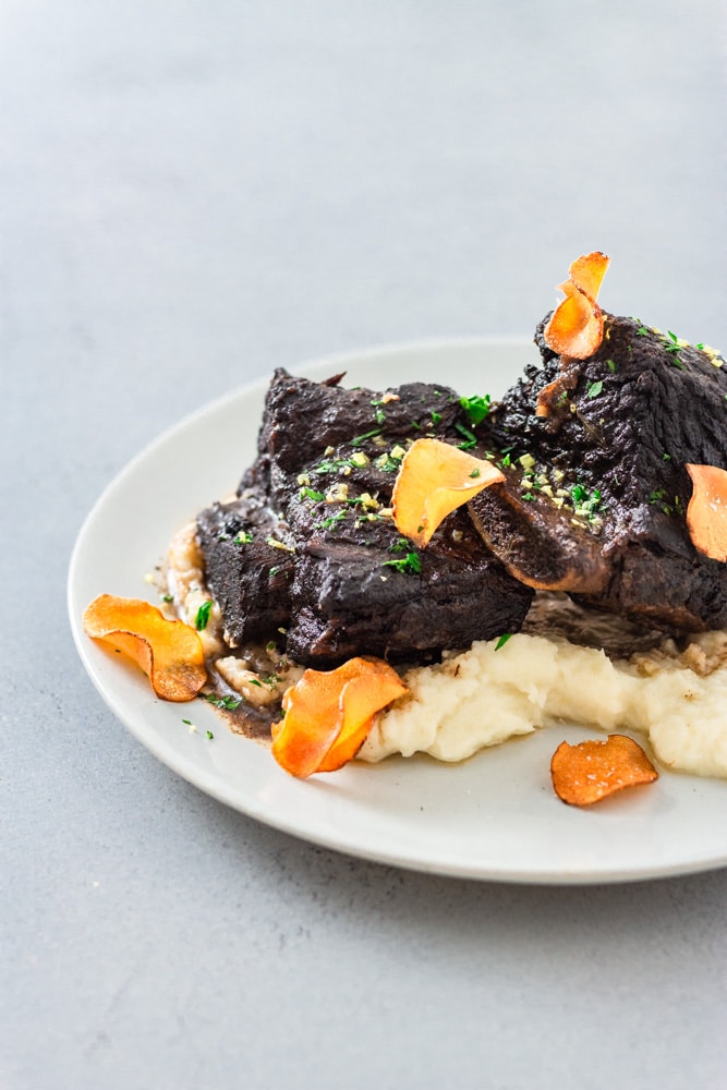 ¾ angled view of a plate of  Braised Short Ribs over sunchoke puree, topped with sunchoke chips, parsley and lemon zest. The surface is light blue.