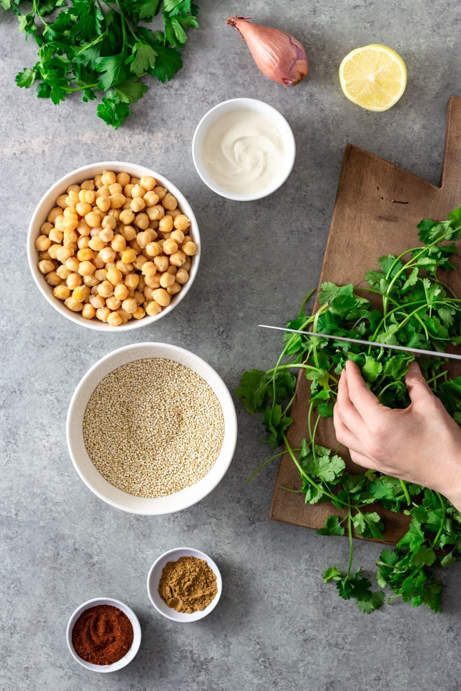 Overhead view of prep for a salad. Cilantro is being cut on a wooden cutting board. There is fresh parsley, a shallot, half of a lemon, a bowl of tahini yogurt, a bowl of chickpeas, a bowl of raw quinoa, and bowls of cumin and paprika on a light grey textured surface. 