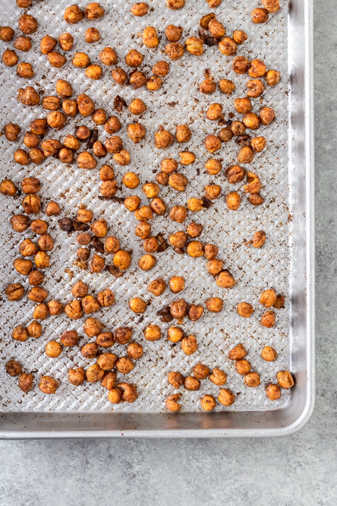 Overhead view of spiced, crispy roasted chickpeas on a baking sheet on a light grey textured surface.