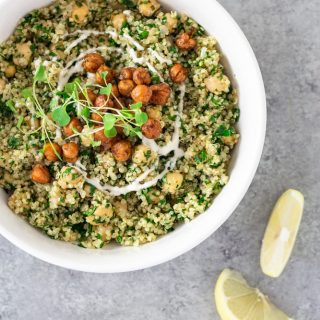 Overhead view of a bowl of Quinoa and Chickpea Salad topped with crispy chickpeas and tahini yogurt, surrounded by lemon wedges, and salad ingredients on a light grey textured surface.