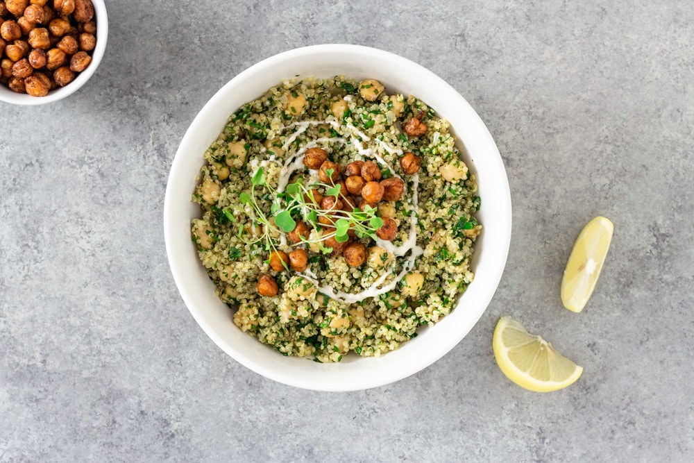 Overhead view of a bowl of Herbed Chickpea and Quinoa Salad with Tahini Yogurt topped with spiced, crispy chickpeas and microgreens, surrounded by lemon wedges and a bowl of crispy chickpeas on a light grey textured surface.