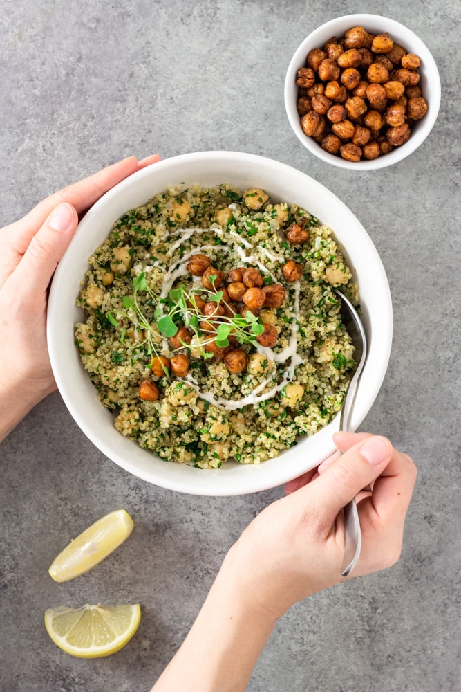 Overhead view of hands sticking a spoon into a bowl of Herbed Chickpea and Quinoa Salad with Tahini Yogurt topped with spiced, crispy chickpeas and microgreens, surrounded by lemon wedges and a bowl of crispy chickpeas on a light grey textured surface.