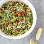 Overhead view of a bowl of Herbed Chickpea and Quinoa Salad with Tahini Yogurt topped with spiced, crispy chickpeas and microgreens, surrounded by lemon wedges on a light grey textured surface.