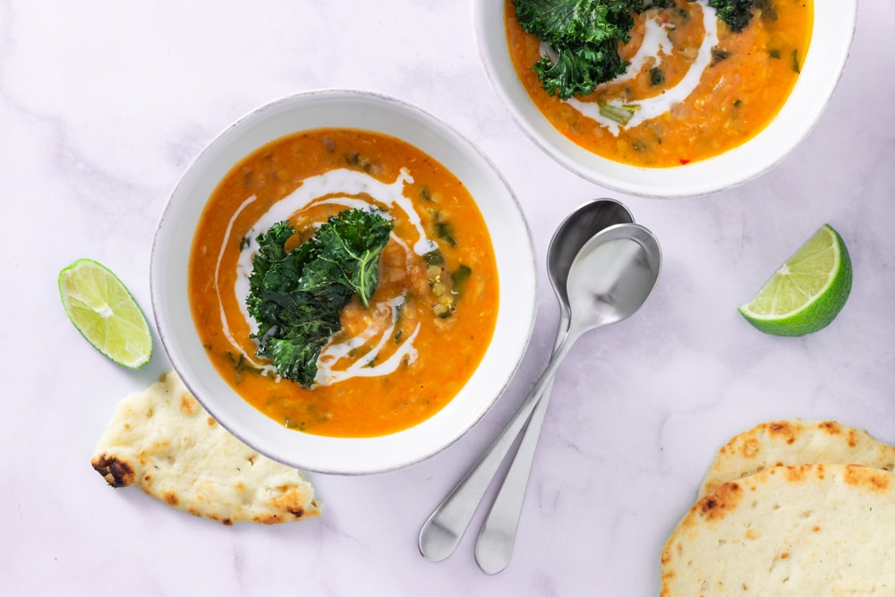 Overhead shot of two bowls of Thai Red Curry Lentil Soup topped with coconut milk and kale chips on a marble surface surrounded by lime wedges, naan bread and spoons.