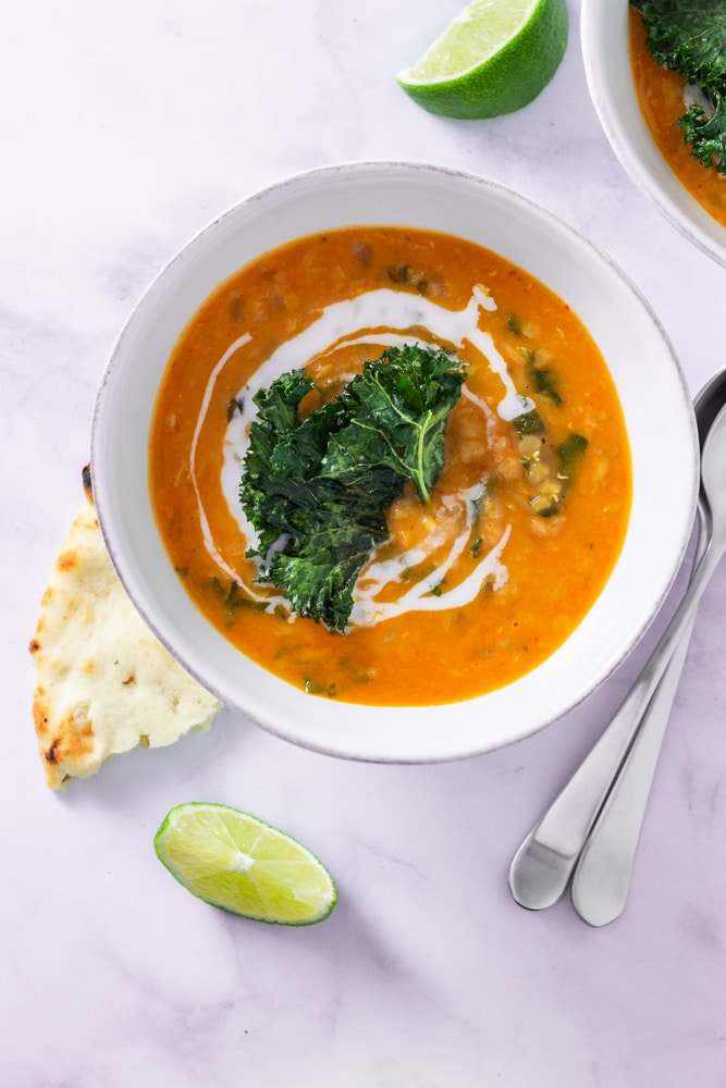 Overhead shot of a bowl of Thai Red Curry Lentil Soup topped with coconut milk and kale chips on a marble surface surrounded by lime wedges, naan bread and spoons.