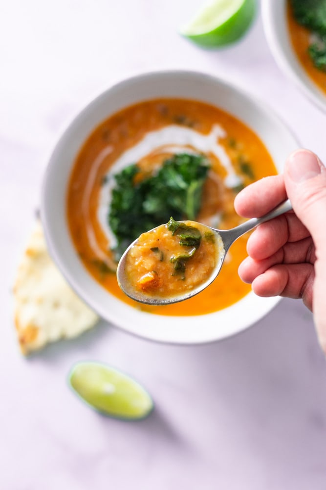 Overhead shot of a spoonful of Thai Red Curry Lentil Soup over a bowl of soup topped with coconut milk and kale chips on a marble surface surrounded by lime wedges, naan bread and spoons.