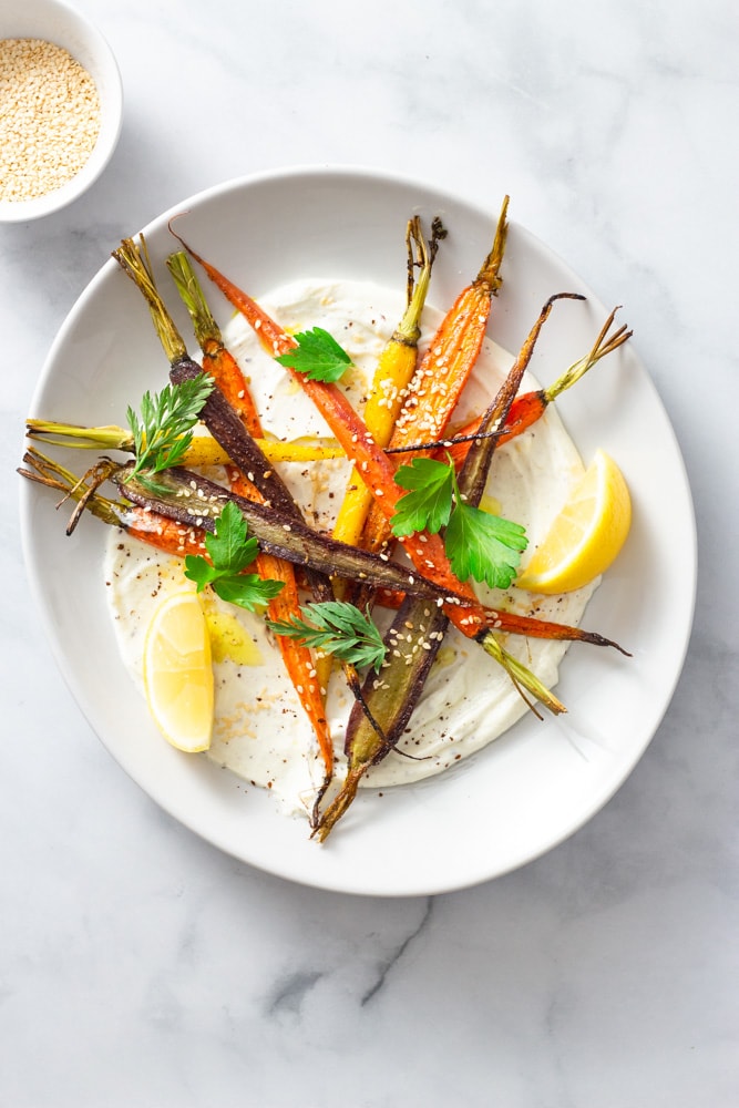 Overhead shot of a plate of sumac yogurt topped with mixed roasted heirloom carrots, topped with parsley and carrot top leaves, sesame seeds and sumac with lemon wedges on a marble surface.