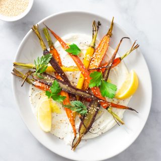 Overhead shot of a plate of sumac yogurt topped with mixed roasted heirloom carrots, topped with parsley and carrot top leaves, sesame seeds and sumac with lemon wedges on a marble surface.