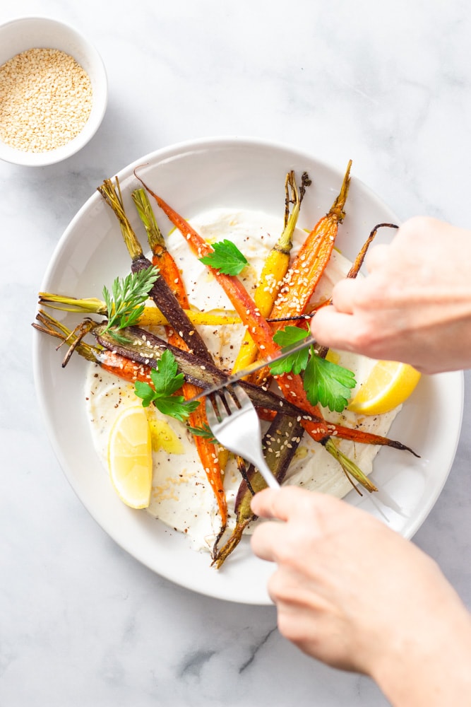 Overhead shot of hands cutting into a plate of sumac yogurt topped with mixed roasted heirloom carrots, topped with parsley and carrot top leaves, sesame seeds and sumac with lemon wedges on a marble surface.