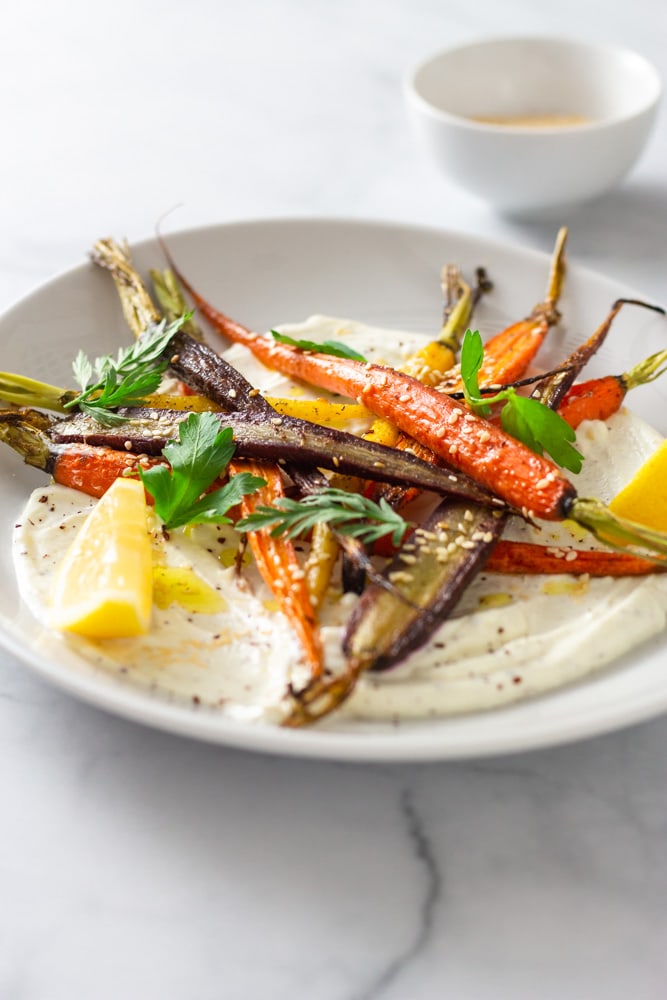¾ angled shot of a plate of sumac yogurt topped with mixed roasted heirloom carrots, topped with parsley and carrot top leaves, sesame seeds and sumac with lemon wedges on a marble surface.