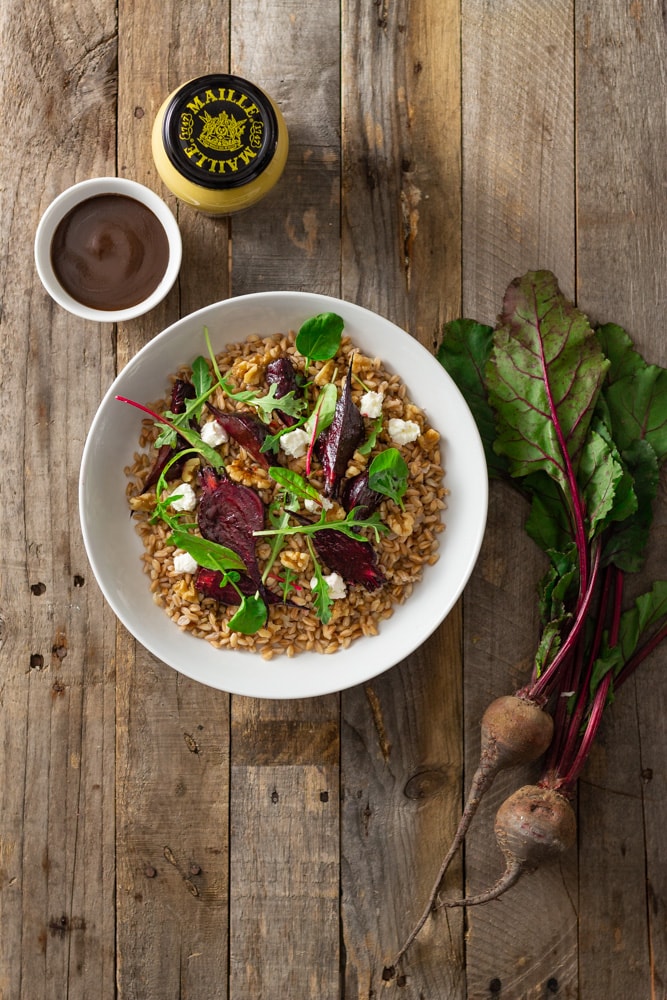 Overhead shot of a dish of farro topped with roasted beets, walnuts, goat cheese, and mixed greens (arugula, watercress, baby chard) with dijon balsamic vinaigrette surrounded by fresh beets with greens, a small bowl of dressing and a jar of Maille mustard on a rustic, grey wood surface.