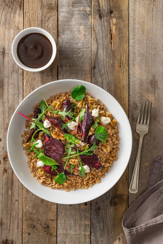 Overhead shot of a dish of farro topped with roasted beets, walnuts, goat cheese, and mixed greens (arugula, watercress, baby chard) with dijon balsamic vinaigrette surrounded by a small bowl of dressing and a rustic silver fork on a rustic, grey wood surface.