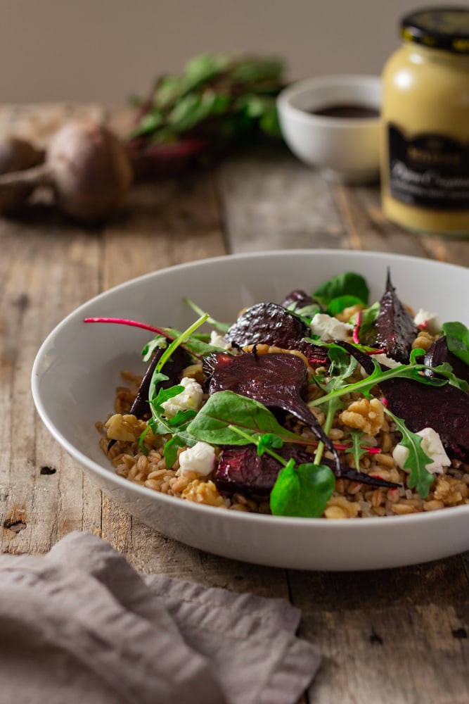 ¾ angled shot of a dish of farro topped with roasted beets, walnuts, goat cheese, and mixed greens (arugula, watercress, baby chard) with dijon balsamic vinaigrette next to a grey dish towel with a small bowl of dressing, fresh beets and a jar of Maille mustard in the background on a rustic, grey wood surface.