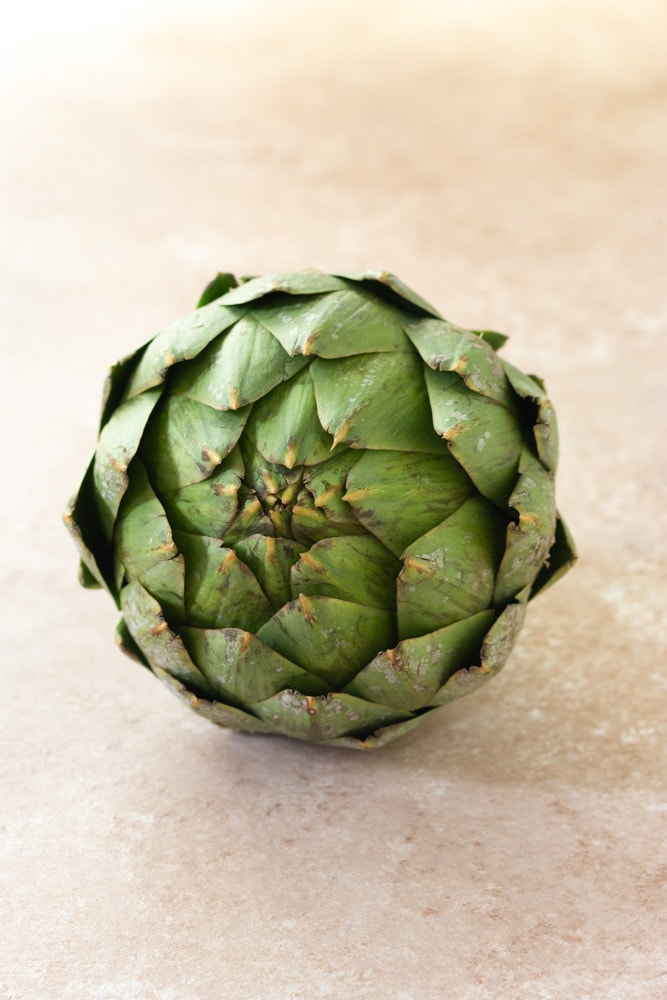 Straight on shot of a raw globe artichoke on a beige textured surface.

