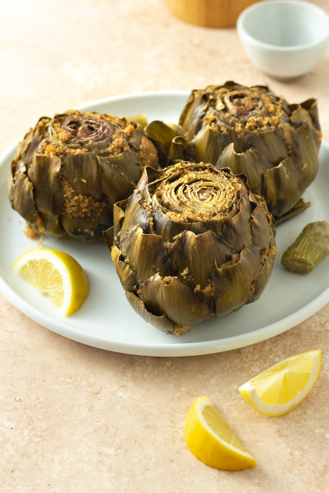 ¾ angled view of Italian Stuffed Artichokes filled with breadcrumbs, cheese, garlic and parsley on a white plate surrounded by lemon wedges, and bowls of salt and pepper on a beige textured surface.