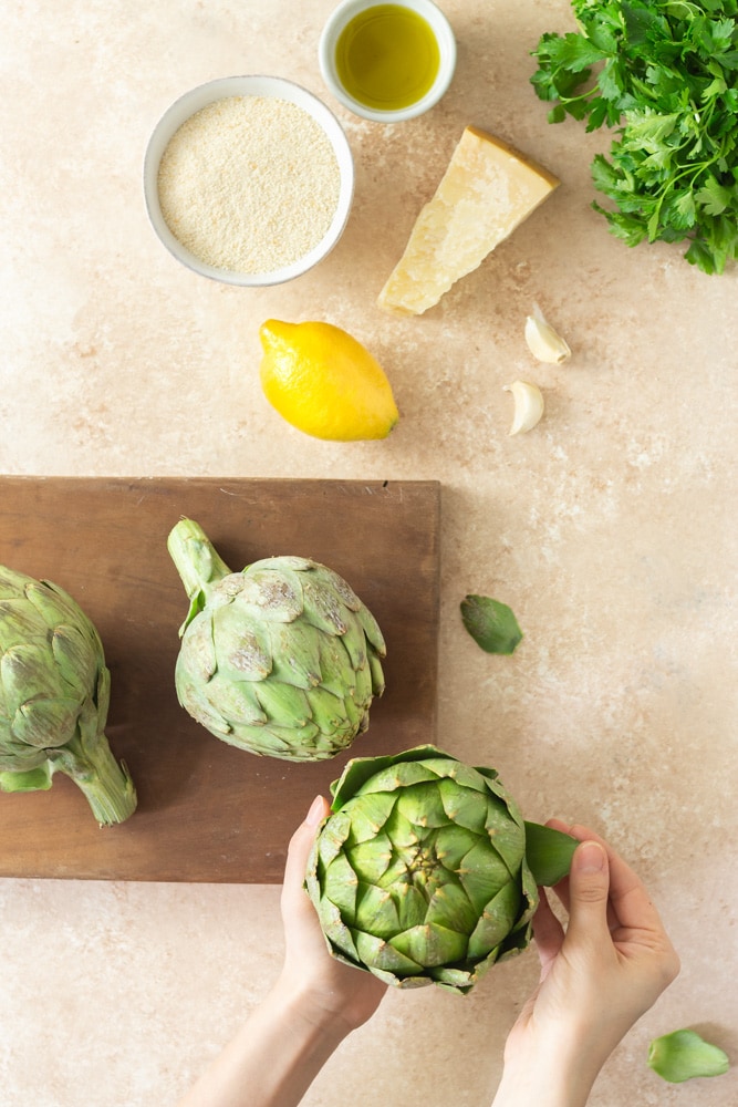 Overhead shot of prep for Italian Stuffed Artichokes. There is parsley, a bowl of olive oil, a bowl of breadcrumbs, a lemon, garlic, a dark wooden cutting board with fresh globe artichokes and hands peeling leaves off an artichoke a beige textured surface.