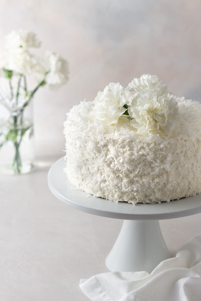 Angled view of a coconut layer cake with Swiss meringue buttercream on a cake stand with a vase of white carnation flowers in the background with a cream colored surface and light textured background.