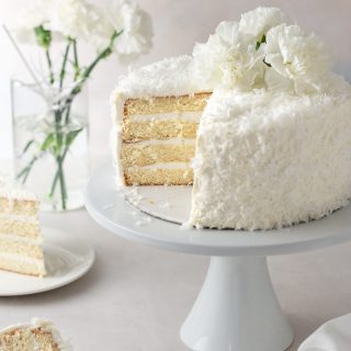 Angled view of a sliced coconut layer cake with Swiss meringue buttercream on a cake stand surrounded by cake slices and a vase of white carnation flowers with a cream colored surface and light textured background.