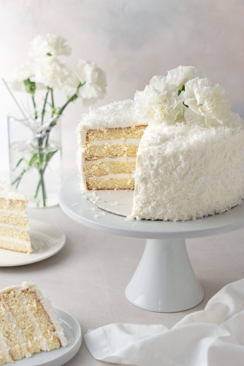 Angled view of a sliced coconut layer cake with Swiss meringue buttercream on a cake stand surrounded by cake slices and a vase of white carnation flowers with a cream colored surface and light textured background.