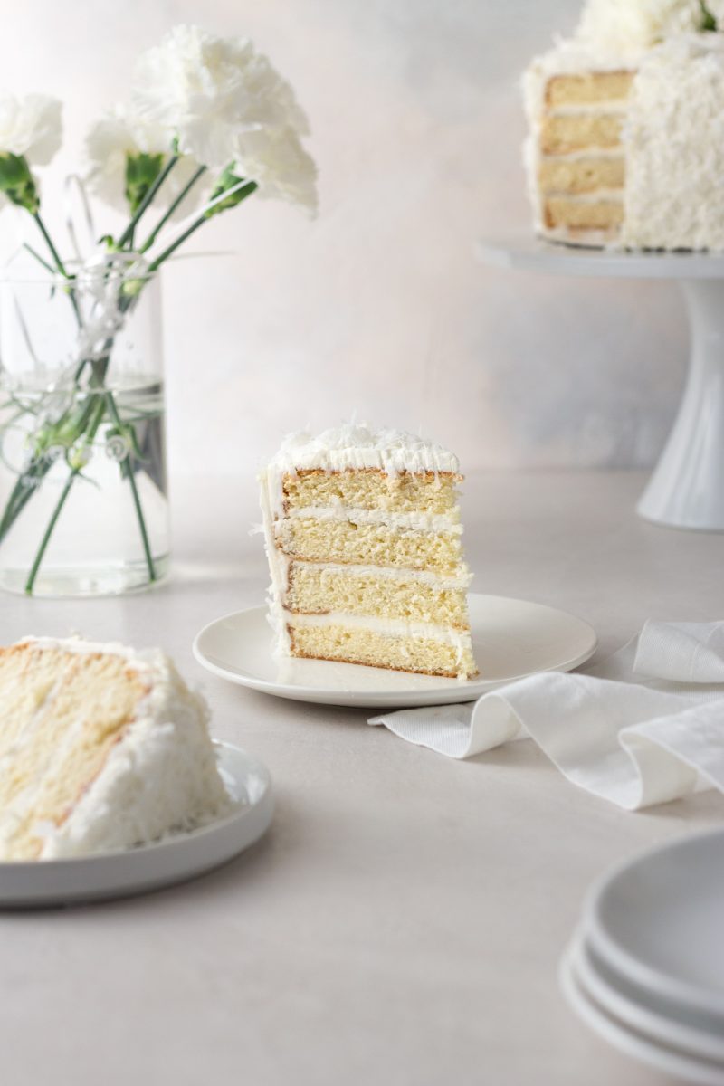 Straight on view of a slice of coconut layer cake with Swiss meringue buttercream on a plate surrounded by another slice of cake, plates, a napkin, a vase of white carnation flowers and the cake on a cake stand with a cream colored surface and light textured background.